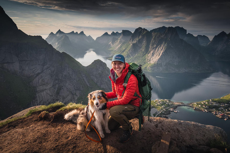 A hiker and a dog posing in front of the dramatic mountains on the top of Reinebringen, Lofoten Islands, Norway