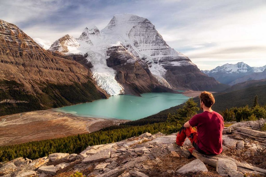 A hiker in a red shirt and red trousers is sitting on a rock with a face directed at a view in front of him. In the distance is snow covered Mount Robson with two glaciers coming down into the turquoise Berg lake. Guide to the Berg Lake Trail 