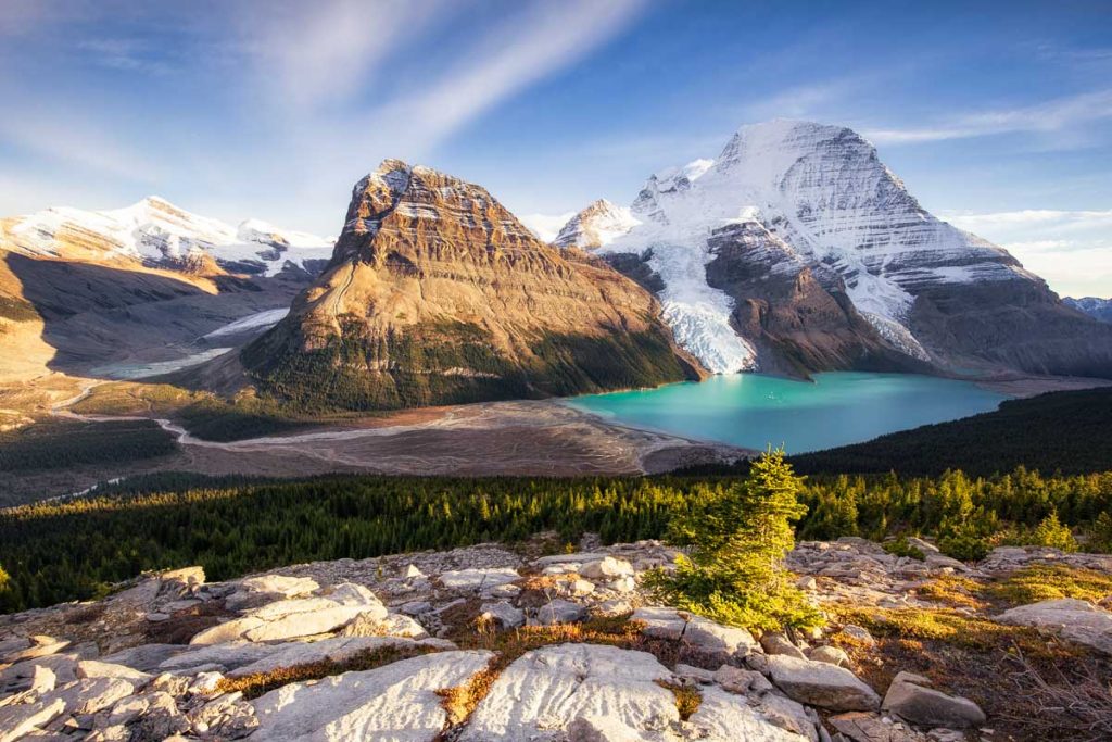 An Aerial view of Mount Robson and Berg Lake. The sun is low in the sky casting beautiful light on the mountains. The lake has an intense turquoise colour. White wispy clouds are covering the blue sky. 