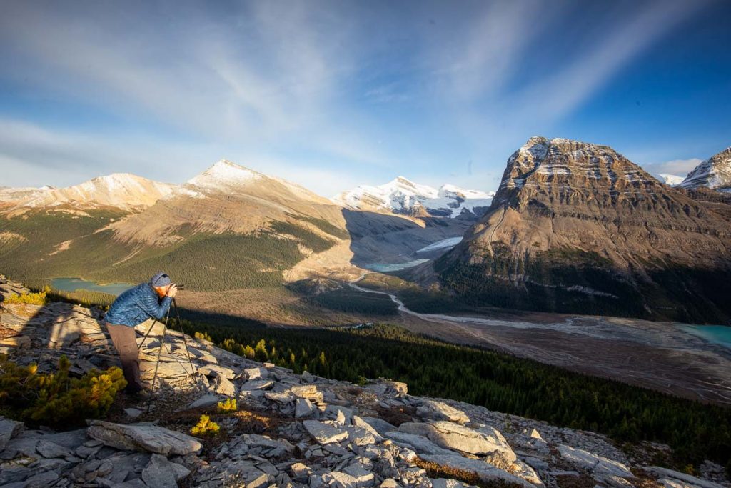 A photographer is bending down to check the scene through the camera viewfinder. The camera is set on a tripod. Behind the photographer is the view down into a valley surrounded by mountains. filled with trees and a river bed flowing from the Robson glacier into Berg lake. Whispy white clouds are covering blue skies. 