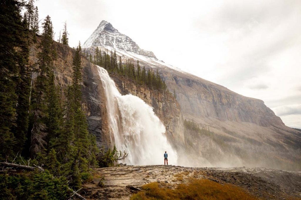 A raging waterfall flowing into the valley below, with pyramid shape peak of Mount Robson peaking right behind it. A hiker is standing in front of the waterfall, looking very small against it. A greyish sky makes the scene a little washed out and gloomy. 